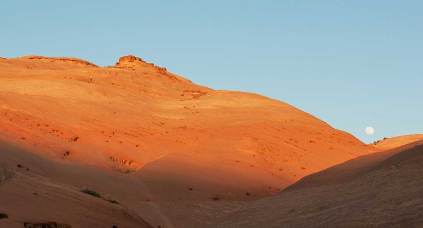 The moon appears above a red mountain ridge in a blue sky